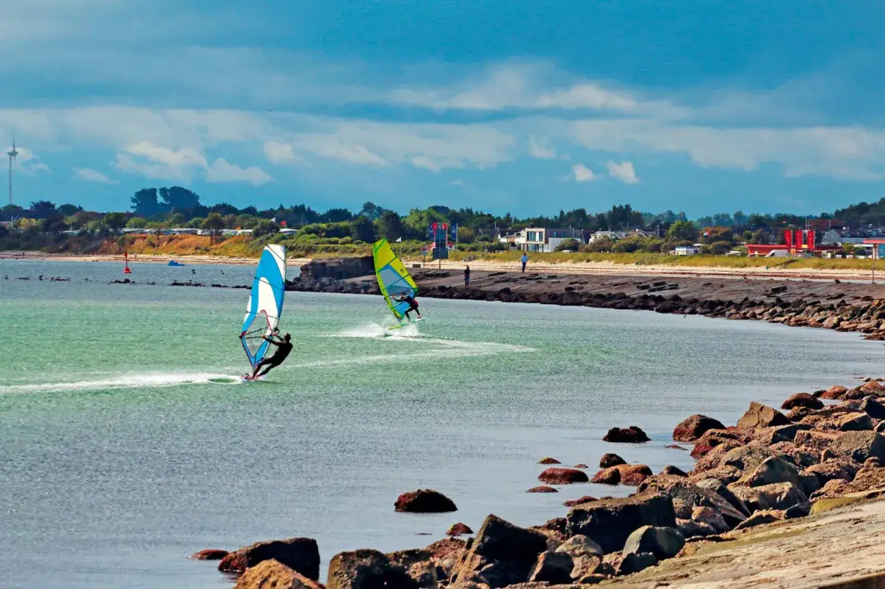 Zwei Windsurfer gleiten &uuml;ber das Wasser. Im Hintergrund eine K&uuml;stenlandschaft mit H&auml;usern und B&auml;umen. Ein typischer Tag f&uuml;r Windsurfen Deutschland.