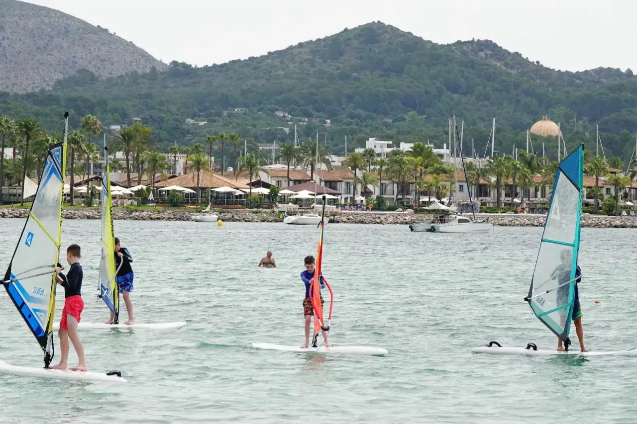 Kinder &uuml;ben Windsurfen auf Mallorca. Im Hintergrund eine K&uuml;stenlinie mit Hotels und Bergen.
