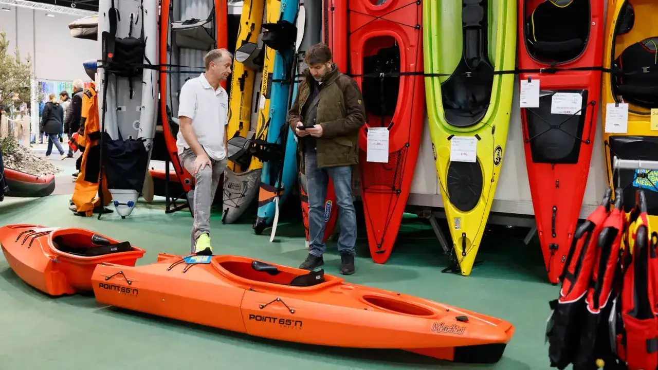 Zwei Männer betrachten Kajaks auf einer Messe. Ein orangefarbenes Kajak steht im Vordergrund. Viele bunte Kajaks sind im Hintergrund ausgestellt, bereit für das nächste Boot Programm.