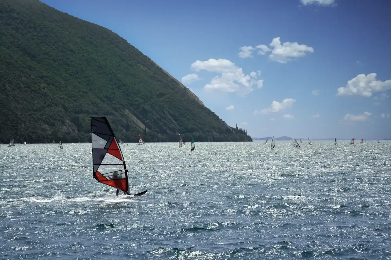 Windsurfer gleitet über glitzerndes Wasser, während am Ufer ein bewaldeter Berg aufragt. Ora wind treibt die Segel.