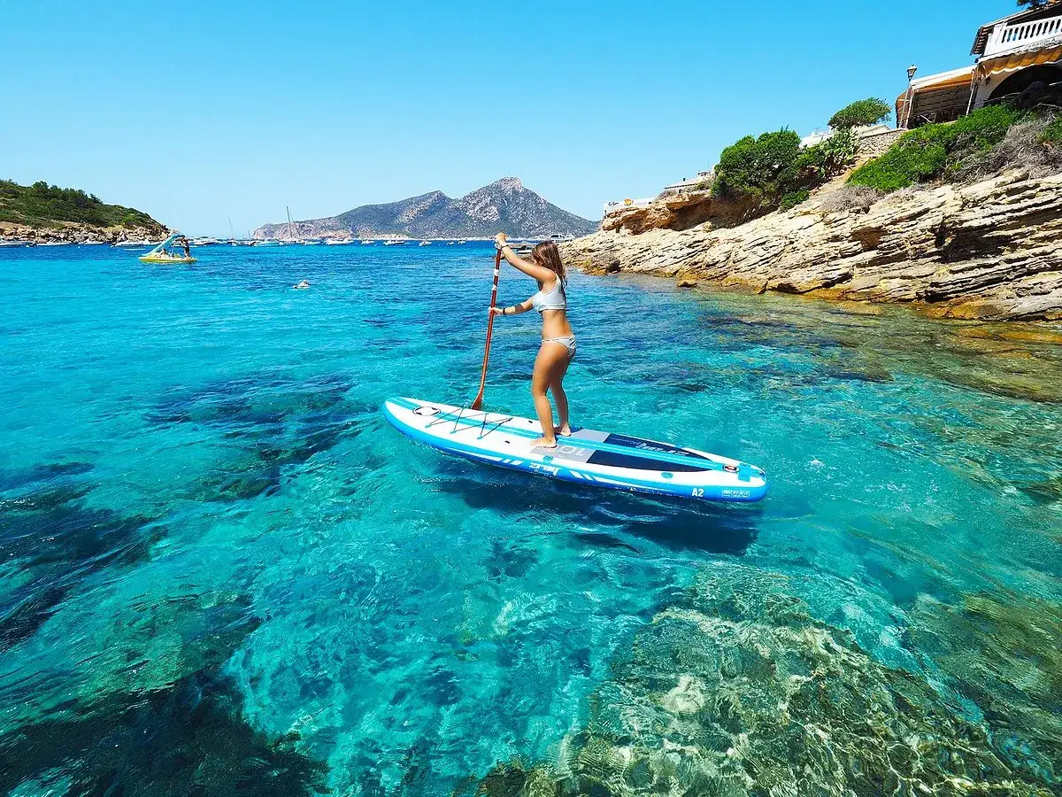 Sup Mallorca: Eine Frau auf einem Stand-Up-Paddleboard genießt das kristallklare, türkisfarbene Wasser vor einer felsigen Küste.