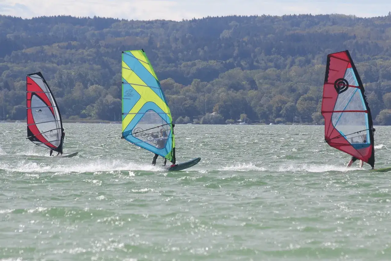 Drei Windsurfer gleiten über den Ammersee. Bunte Segel im Wind, im Hintergrund bewaldete Hügel.