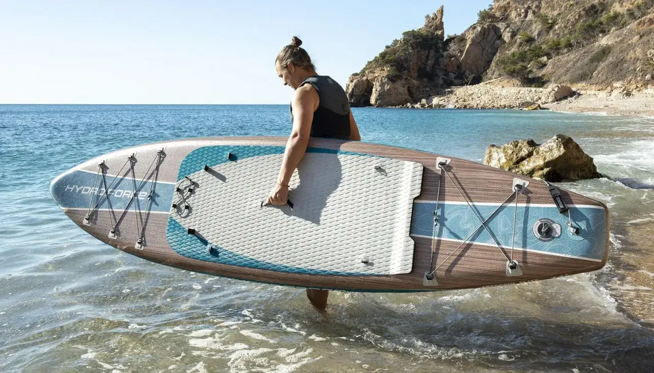 Mann mit SUP-Board im Wasser, bereit für SUP Training. Sonnige Bucht mit Felsen im Hintergrund.