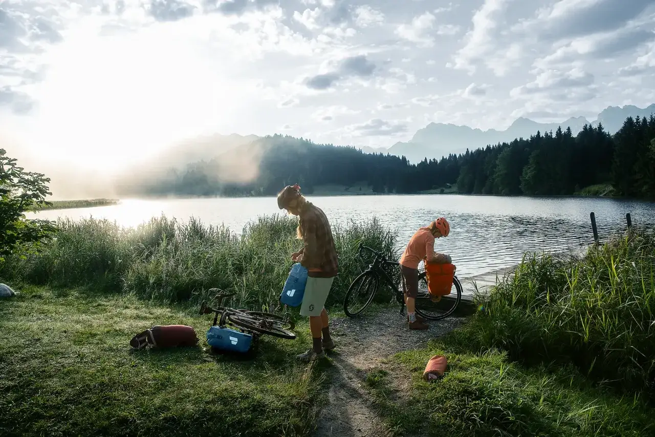 Zwei Radreisende packen ihre Ausrüstung am See. Einer reinigt seinen Rucksack, während die Sonne über den Bergen aufgeht.