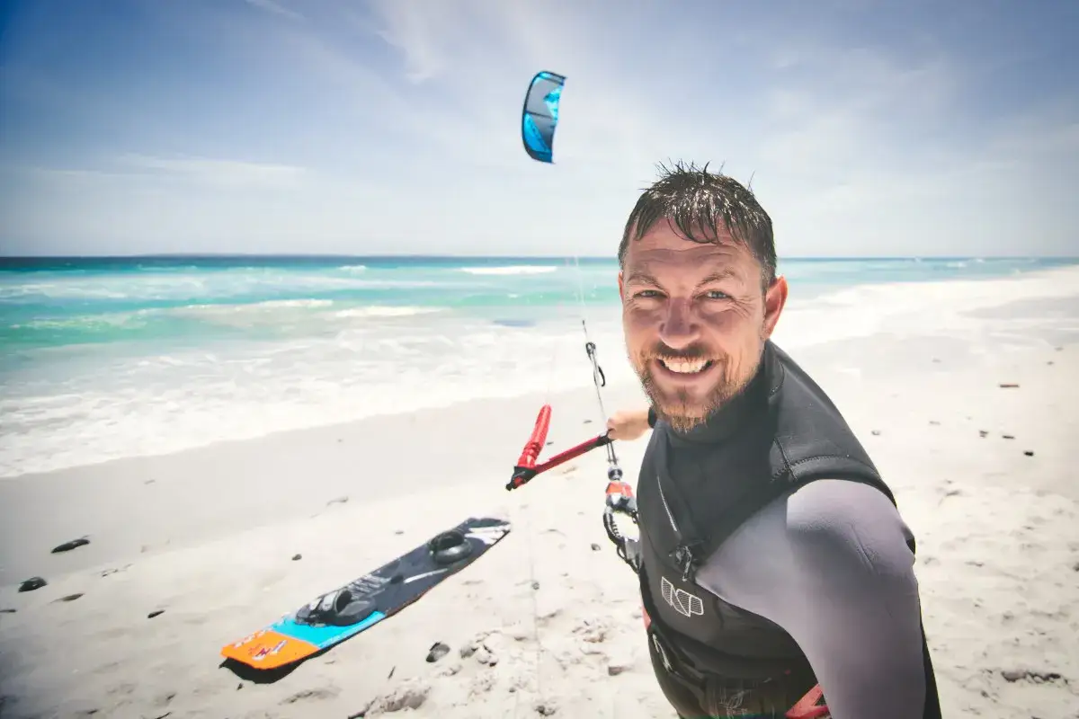 Ein Mann mit einem breiten L&auml;cheln steht am Strand, bereit zum surfen mit drachen in der hand. Ein Kiteboard liegt im Sand, ein blauer Kite schwebt am Himmel.