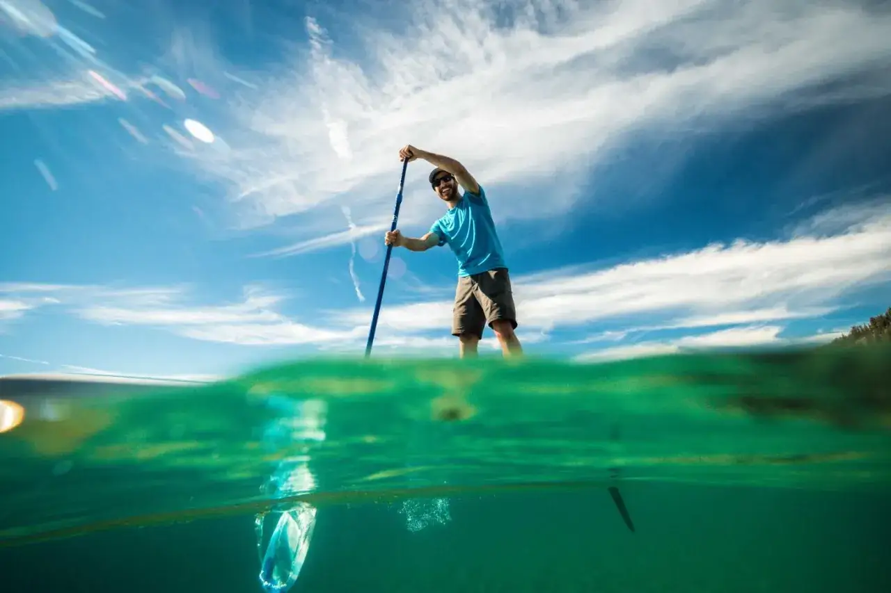 Mann auf SUP-Board, der geradeaus fährt, mit blauem Himmel und Wolken. Unterwasseraufnahme zeigt grünes Wasser.