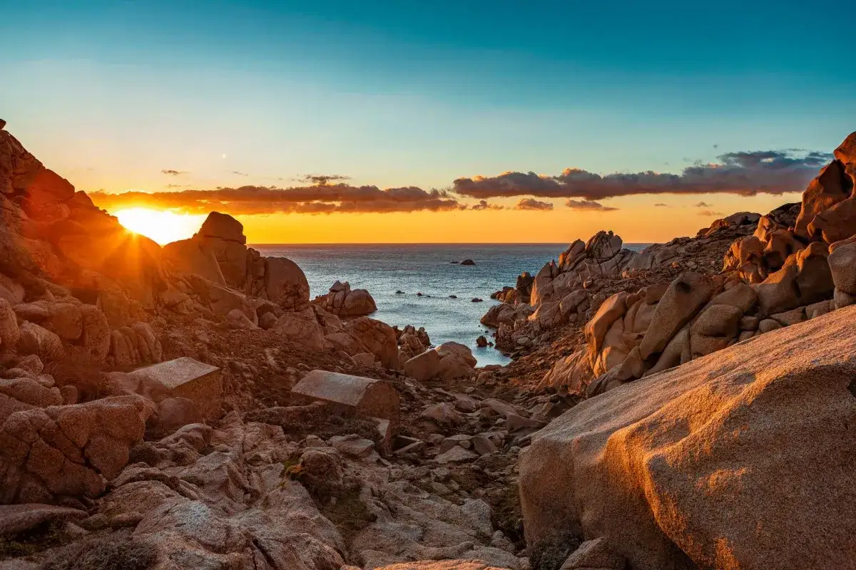 Sonnenuntergang am Capo Testa. Felsige K&uuml;ste mit kleinen Steinh&auml;usern, das Meer glitzert im warmen Licht.