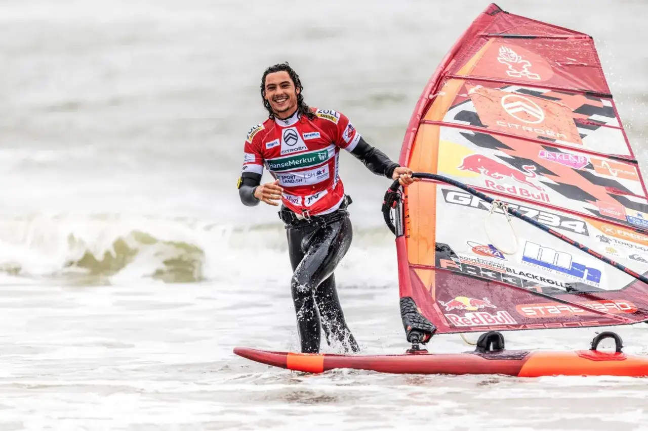 Bekannte deutsche Windsurfer: Ein lachender Mann im Neoprenanzug mit seinem roten Segel im Wasser.