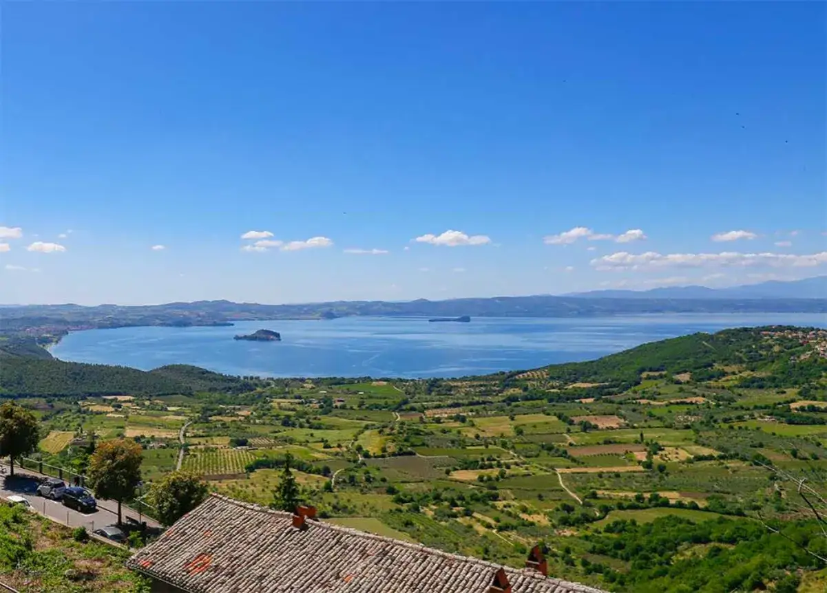 Blick &uuml;ber das Lago di Bolsena mit gr&uuml;nen H&uuml;geln, Feldern und einer Insel im Wasser unter blauem Himmel.