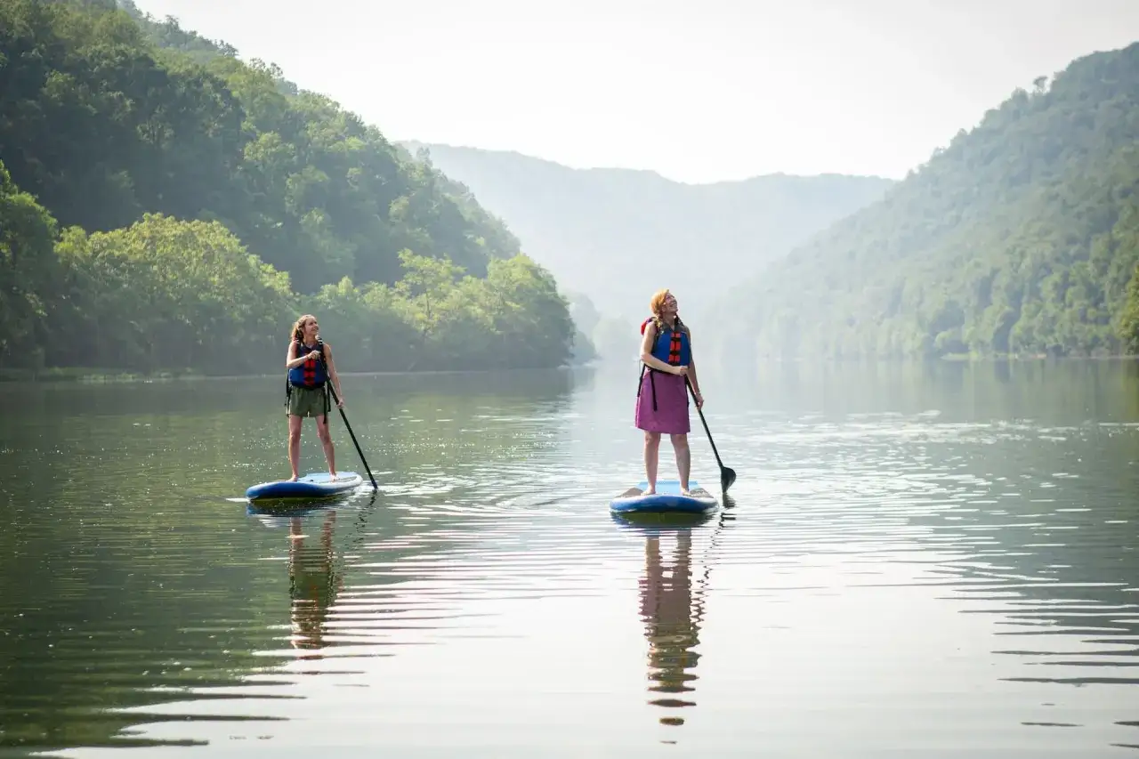 Zwei Frauen genie&szlig;en eine entspannte SUP-Tour auf einem ruhigen Fluss, umgeben von sanften, bewaldeten H&uuml;geln.
