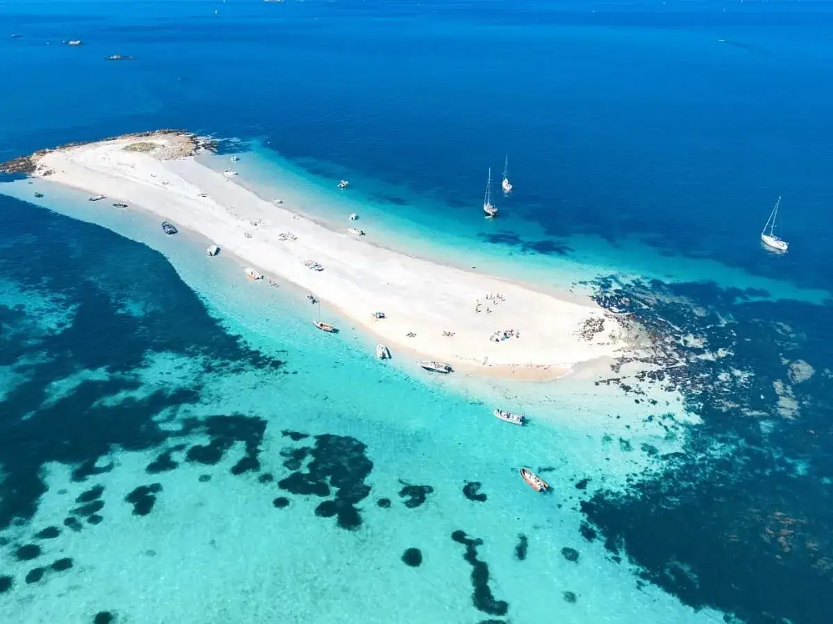 Traumhafte wei&szlig;e Sandb&auml;nke und t&uuml;rkisfarbenes Wasser auf den Glenan Inseln. Boote ankern in der Bucht, w&auml;hrend Menschen am Strand entspannen.