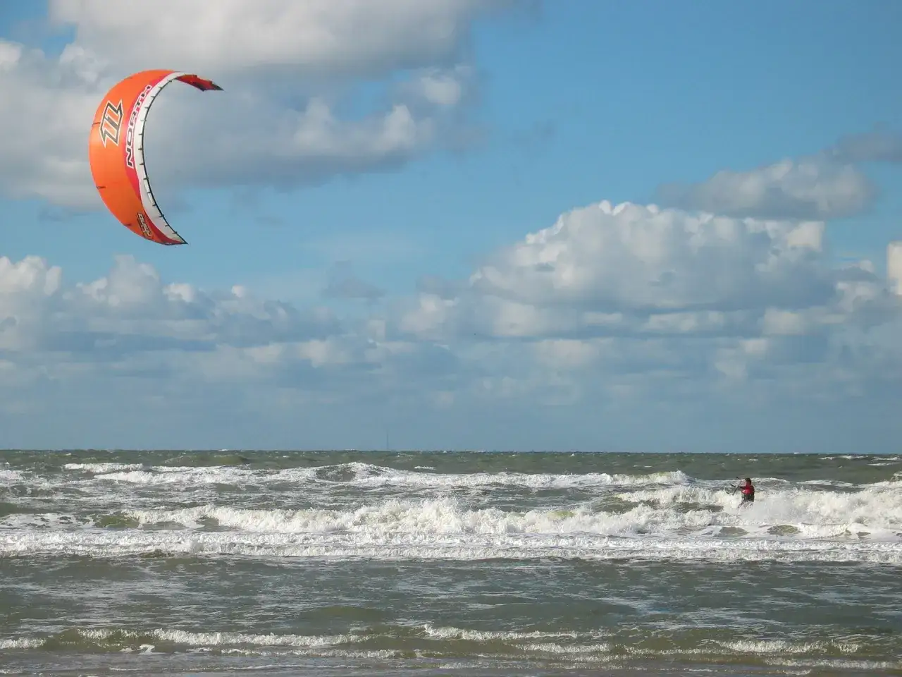 Ein Kiteboarder surft auf den Wellen unter einem orangefarbenen Kite.