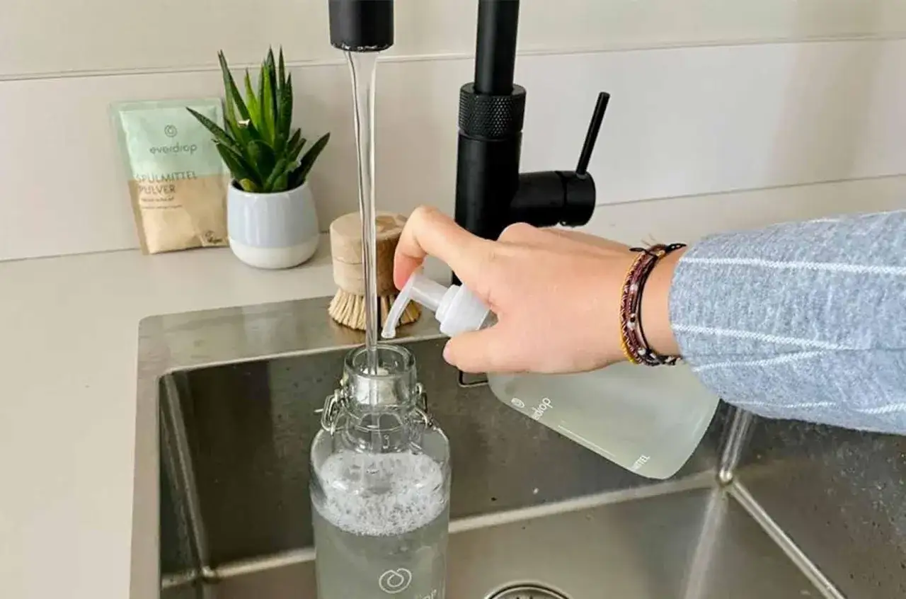 Eine Hand dr&uuml;ckt Sp&uuml;lmittel in eine Glasflasche, w&auml;hrend Wasser aus dem Wasserhahn l&auml;uft. So einfach ist die Trinkflasche reinigen.