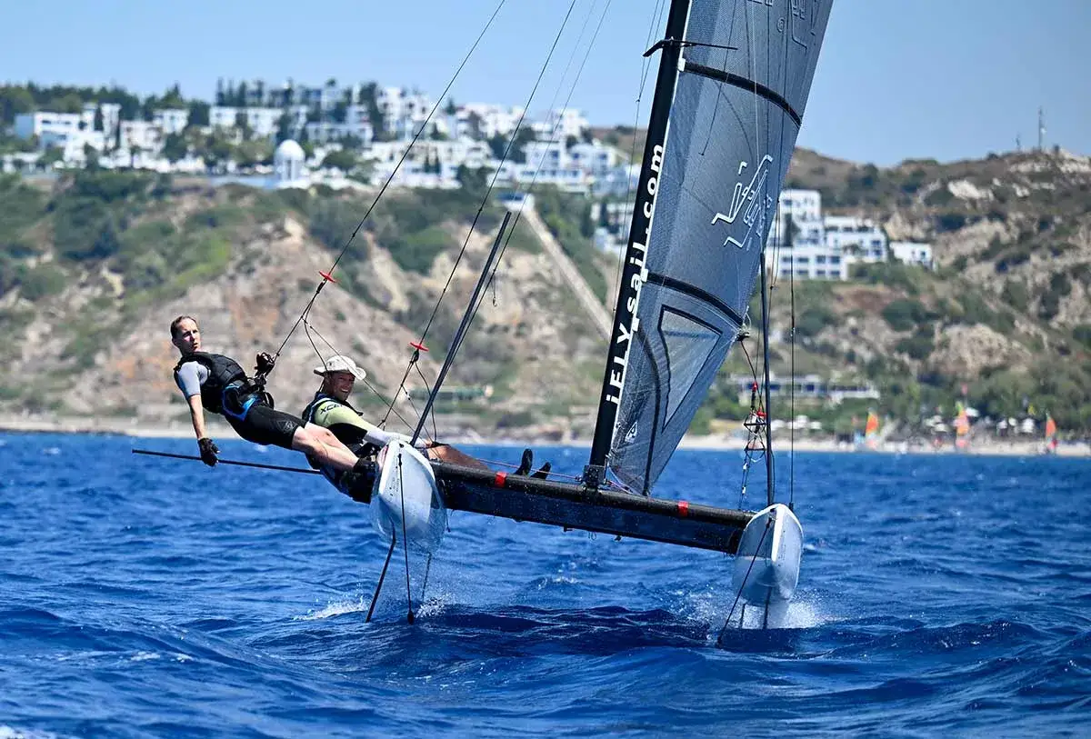 Zwei M&auml;nner auf einem Katamaran, der mit Foil surfen ohne segel &uuml;ber das Meer gleitet. Im Hintergrund eine K&uuml;ste mit wei&szlig;en H&auml;usern.