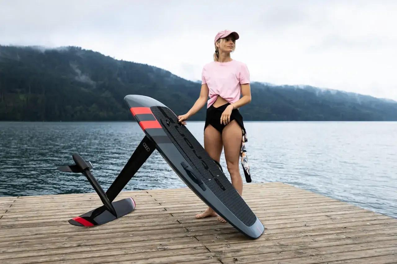 Frau mit pinkem Shirt und Kappe steht neben einem Surfbrett mit Motor auf einem Holzsteg.