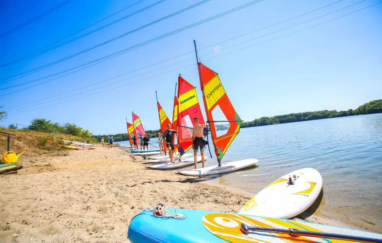 Sonnenklarer Tag am See in NRW: Mehrere Personen bereiten sich auf das Windsurfen vor, bunte Segel stehen am Strand.