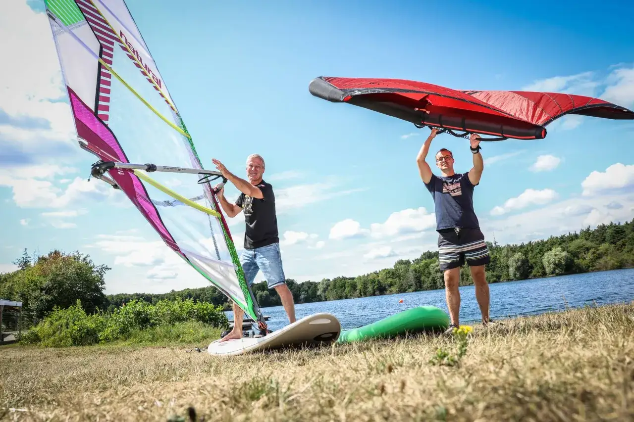 Zwei Männer bereiten sich auf Windsurfen NRW vor. Einer hält ein Segel, der andere einen roten Wing.