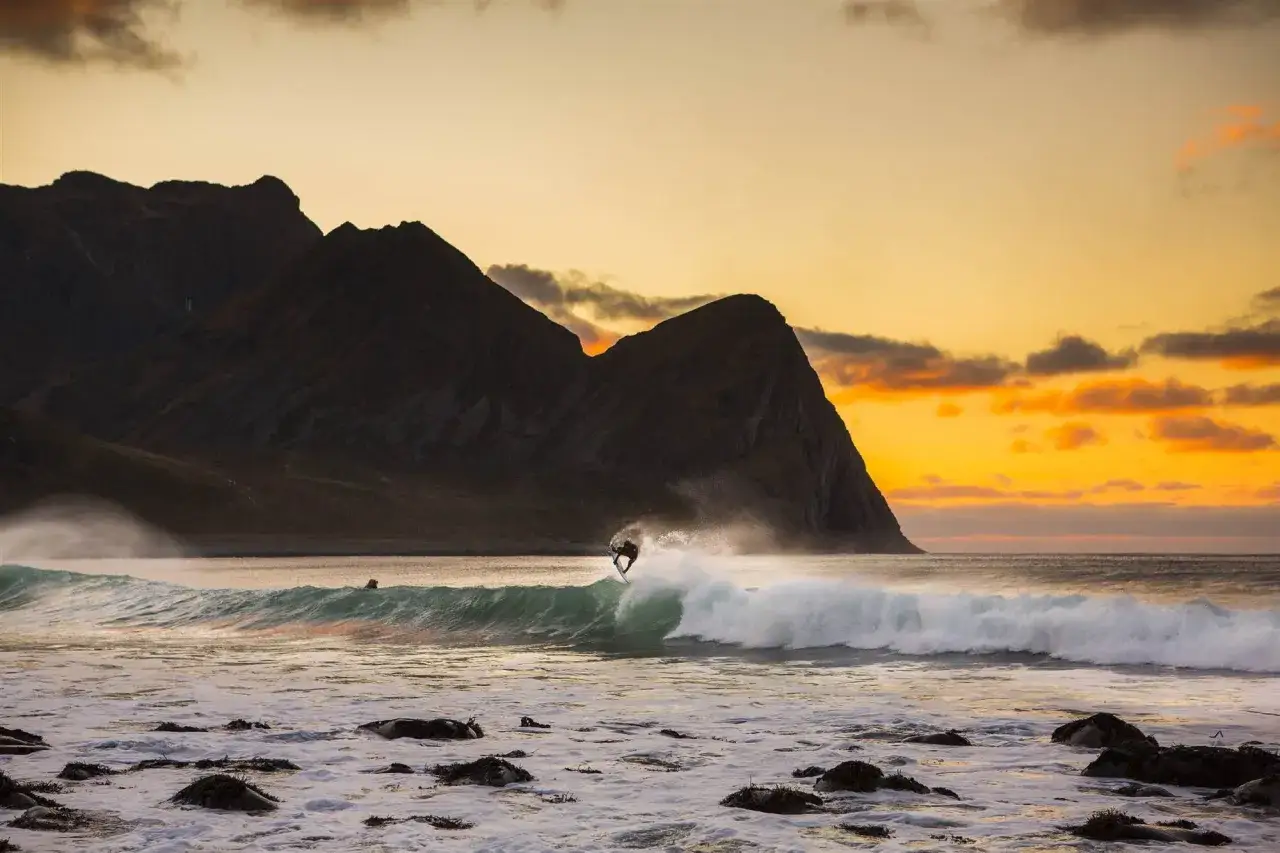 Surfer springt in der Luft auf einer Welle vor dramatischer Bergkulisse bei Sonnenuntergang. Ein atemberaubendes Bild von Norwegen Surfen.
