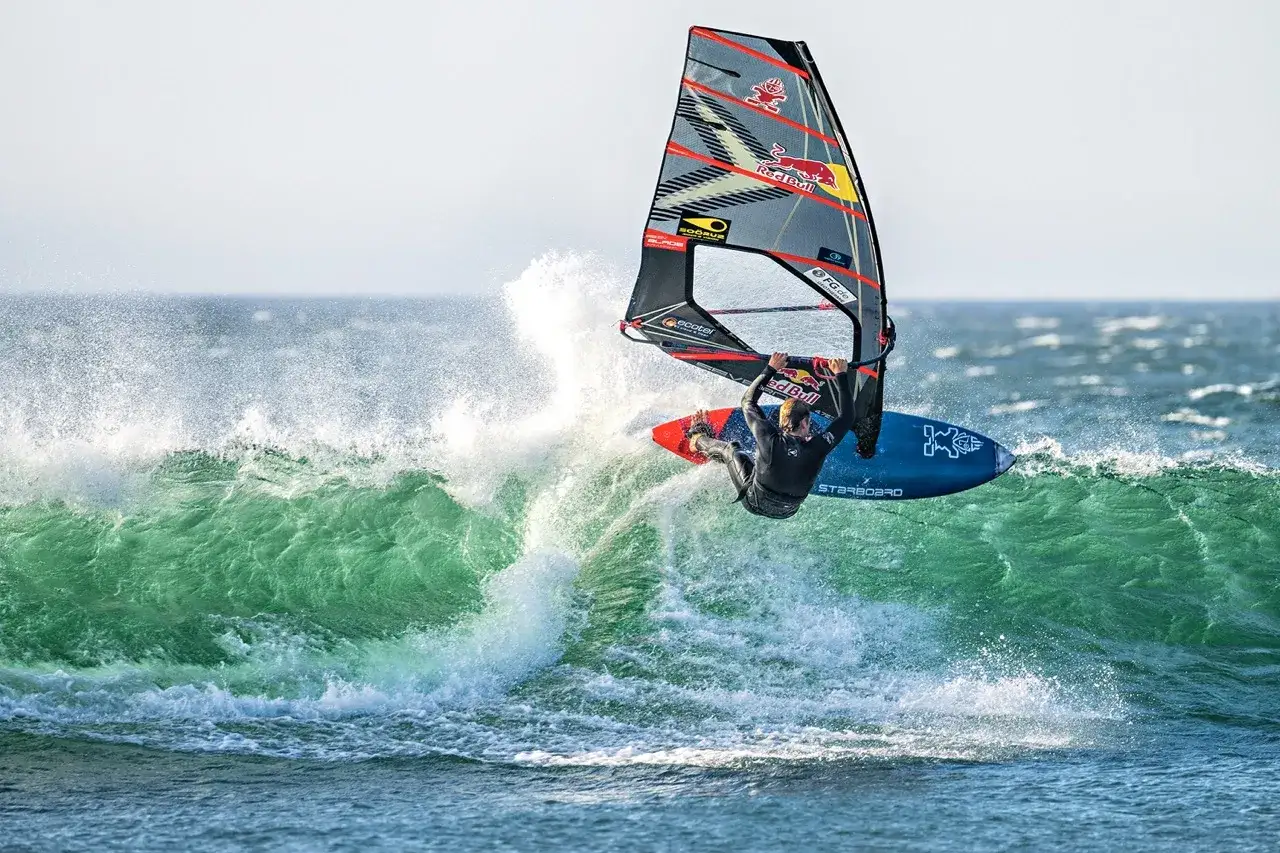 Liam Dunkerbeck fliegt mit seinem Windsurfbrett &uuml;ber eine gr&uuml;ne Welle, die Gischt spritzt.