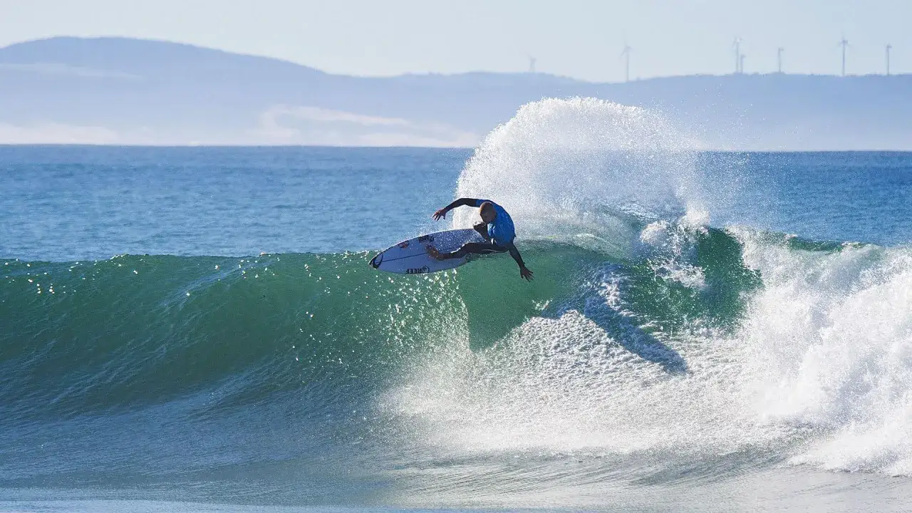 Ein Surfer auf Zypern reitet eine grüne Welle, umgeben von Gischt. Im Hintergrund sind sanfte Hügel mit Windrädern zu sehen.