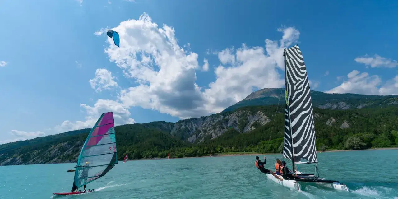 Windsurfen in Frankreich: Ein bunter Segelboot-Mix auf türkisfarbenem Wasser vor bewaldeten Bergen.