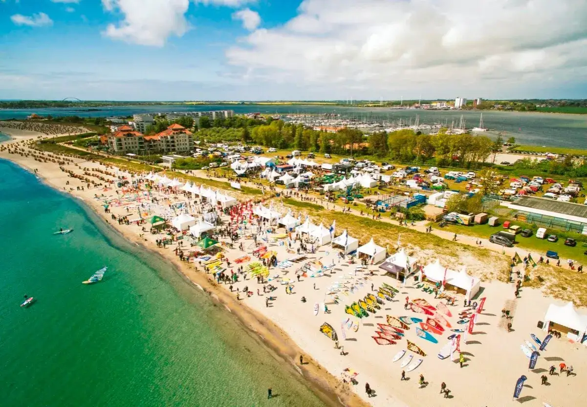 Gro&szlig;e Veranstaltung am Strand mit vielen Zelten, Menschen und Ausr&uuml;stung f&uuml;r Windsurfen Ostsee. Boote im Wasser und eine Marina im Hintergrund.