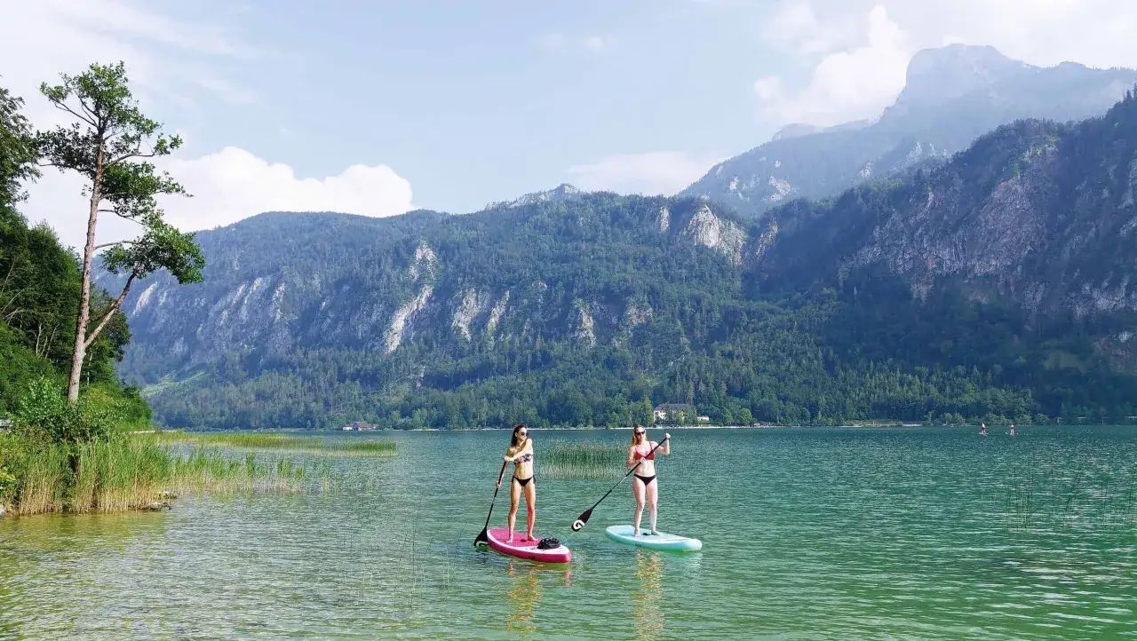 Zwei Frauen genie&szlig;en Stand-Up-Paddling auf einem ruhigen See vor einer Bergkulisse. Perfekte SUP Spots f&uuml;r einen entspannten Tag.
