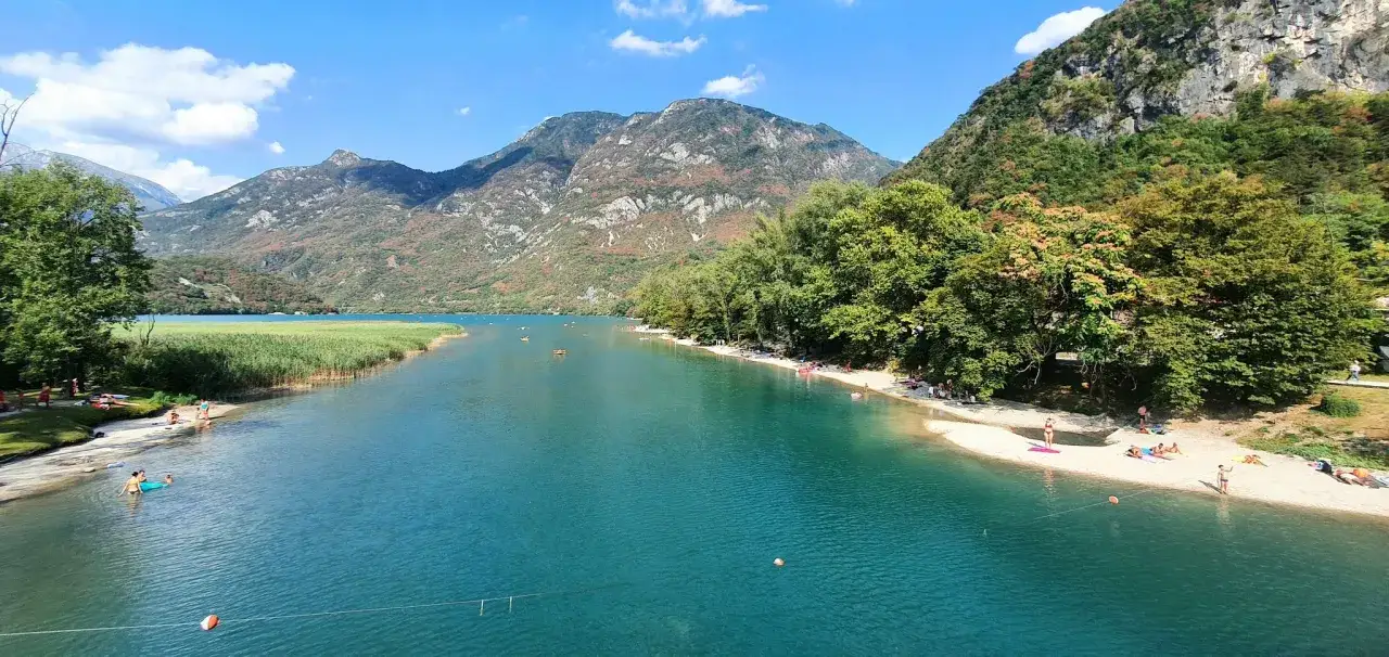 Türkises Wasser am Lago di Cavazzo, umgeben von grünen Hügeln und Bergen. Menschen entspannen am sandigen Ufer und genießen das klare Wasser.