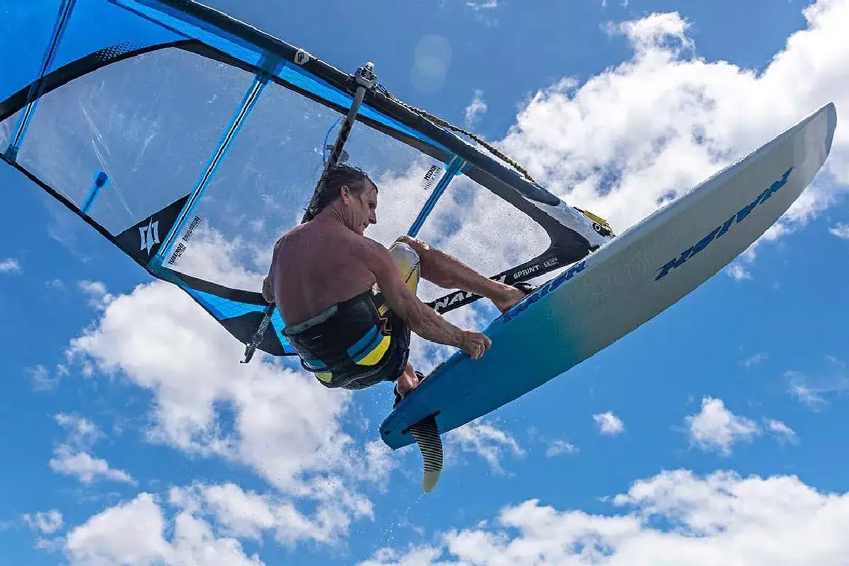 Ein Windsurfer mit einem freeride windsurfboard springt hoch in den blauen Himmel mit Wolken.