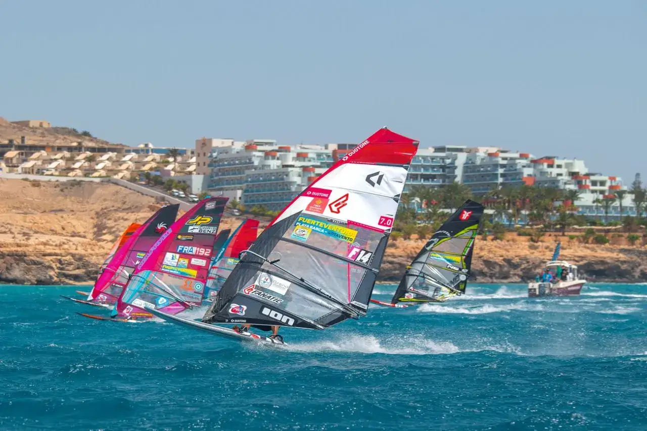 Windsurfer auf Fuerteventura, die Segel sind bunt und die Wellen blau. Im Hintergrund Hotels und eine felsige Küste.