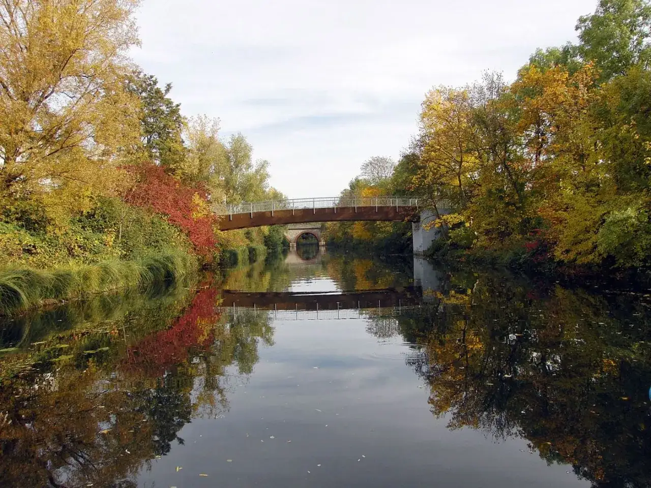 Herbstliche Farben am Fluss in Leipzig. Eine Brücke überspannt das Wasser, das die Bäume und den Himmel spiegelt. Sup Leipzig!