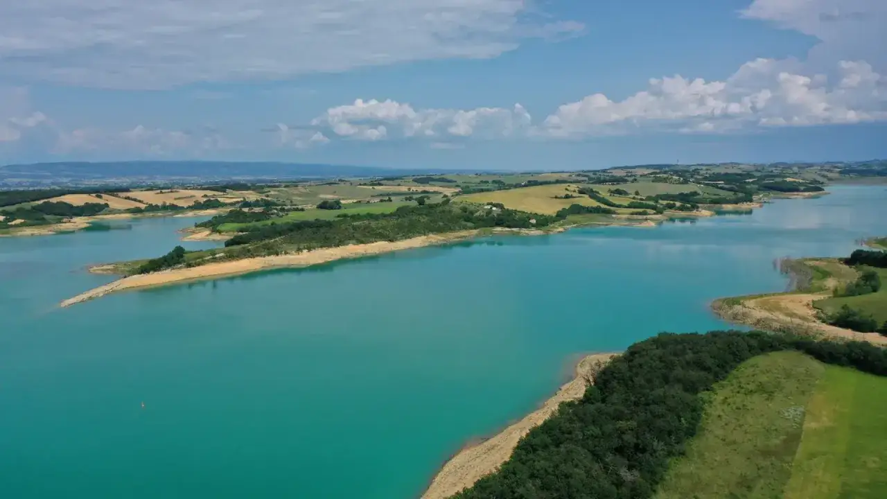 Türkisfarbener Lac de Ganguise, umgeben von grünen Hügeln und Feldern unter einem blauen Himmel mit Wolken.