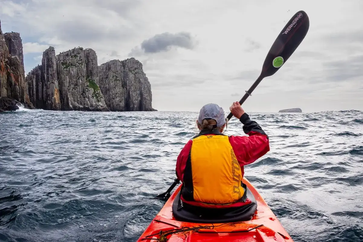 Person im Kajak hält das Kajak Paddel richtig, navigiert auf dem Meer an steilen Felsen vorbei.