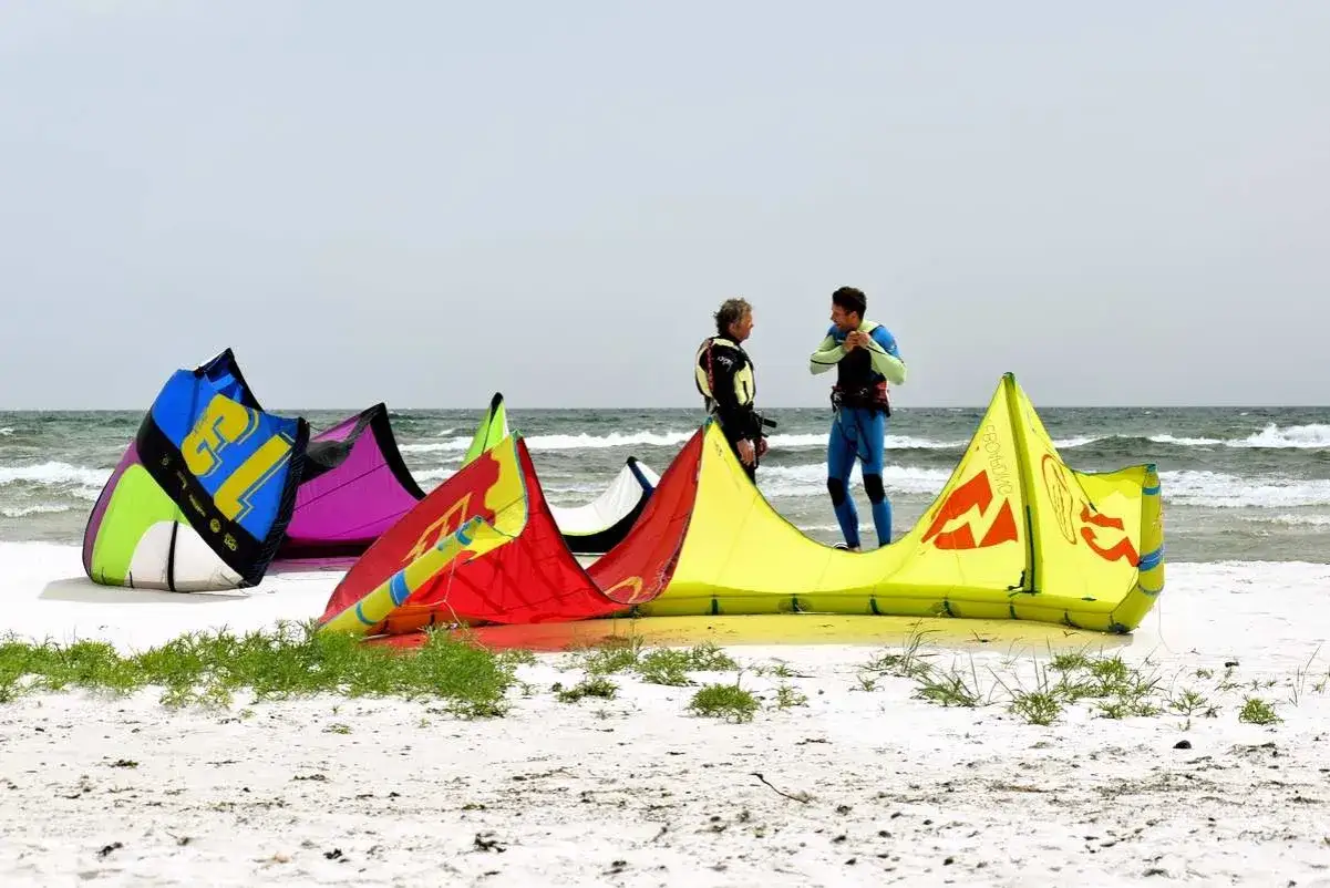 Zwei Männer in Neoprenanzügen bereiten sich auf das Bornholm Surfen vor, bunte Kites liegen am Strand.