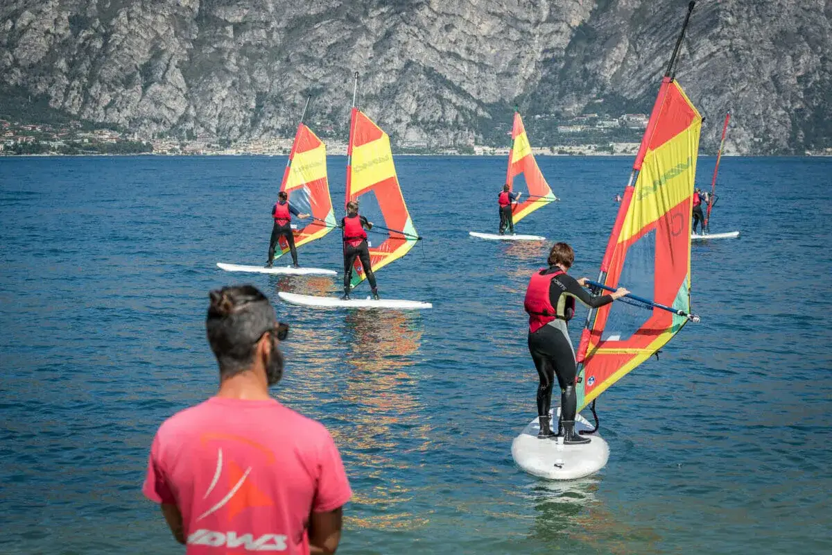 Windsurfer &uuml;ben am Gardasee. Mehrere Personen auf Surfbrettern mit bunten Segeln gleiten &uuml;ber das blaue Wasser vor einer Bergkulisse.