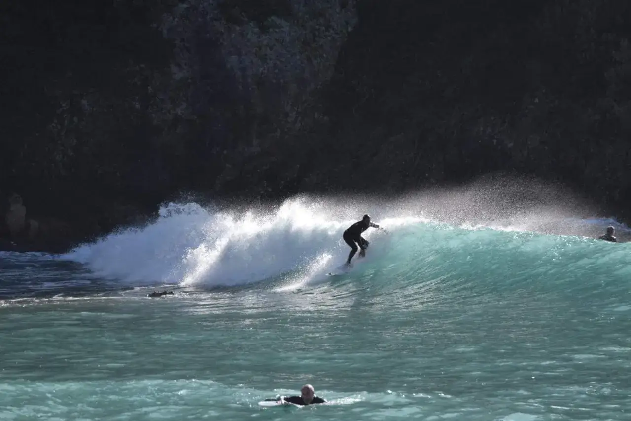 Surfer reitet eine Welle vor dunkler Felswand. Ein weiterer Surfer schwimmt im Vordergrund. Surfen Griechenland.