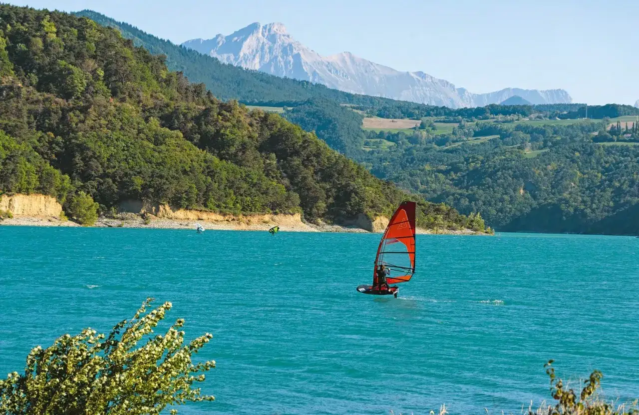 Windsurfer auf dem Lac de Monteynard, umgeben von grünen Hügeln und majestätischen Bergen.