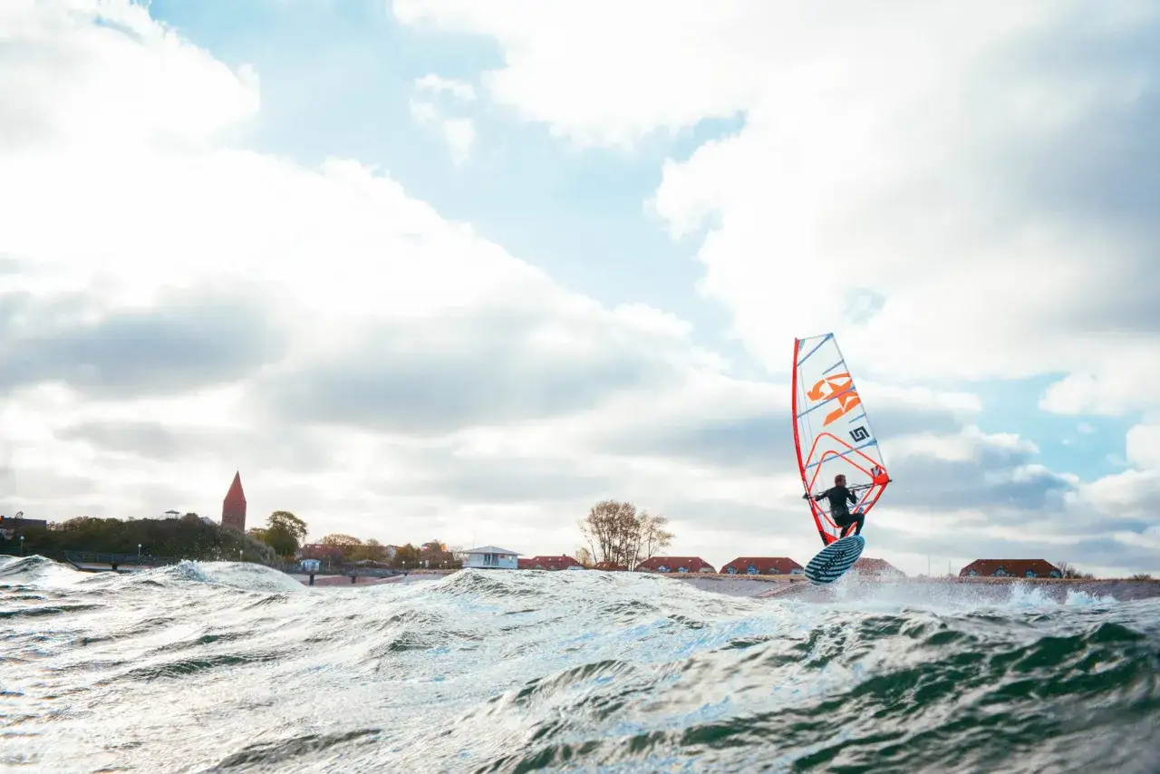 Ein Windsurfer gleitet &uuml;ber die Wellen, das bunte Segel im Wind. Die K&uuml;stenlinie mit H&auml;usern und einem Turm im Hintergrund. Ein toller Tag zum pepelow surfen.