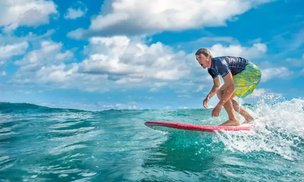 Ein Surfer reitet eine Welle vor der Küste der Dominikanischen Republik. Der Mann trägt ein blaues Shirt und bunte Badeshorts.