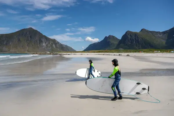 Zwei Surferinnen mit Neoprenanzügen und Surfbrettern am Strand in Norwegen. Die Berge im Hintergrund sind grün und die Wellen sind sanft.
