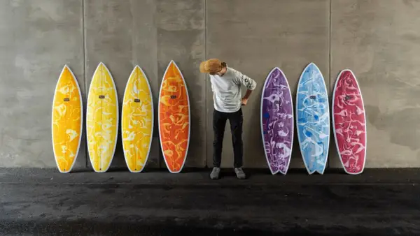 Mann mit Hut steht vor bunten Surfbrettern, bereit für das Wellenreiten.