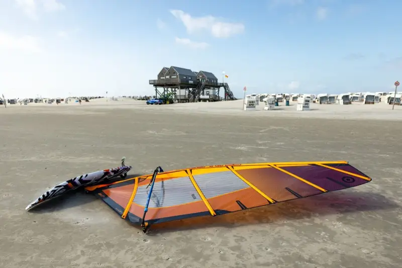 Windsurfen St. Peter-Ording: Segel und Brett liegen am Strand, bereit für den nächsten Ritt auf den Wellen. Strandkörbe und das markante Stelzenhaus im Hintergrund.