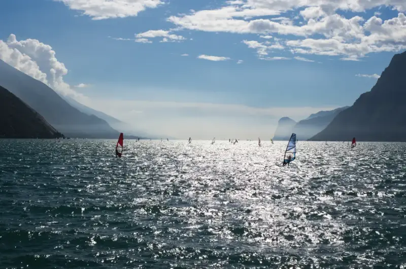 Windsurfer tummeln sich auf dem glitzernden Gardasee vor einer Bergkulisse.