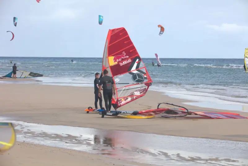 Windsurfer bereiten sich am Strand von Pozo Izquierdo, Gran Canaria, auf das Wasser vor. Mehrere bunte Segel und Boards liegen am Sand.