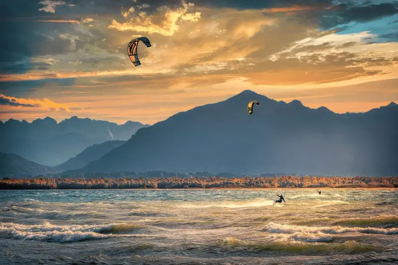 Kitesurfer gleiten über die Wellen am Chiemsee, während die Sonne hinter den Bergen untergeht. Ein traumhafter Abend für Windsurfen.
