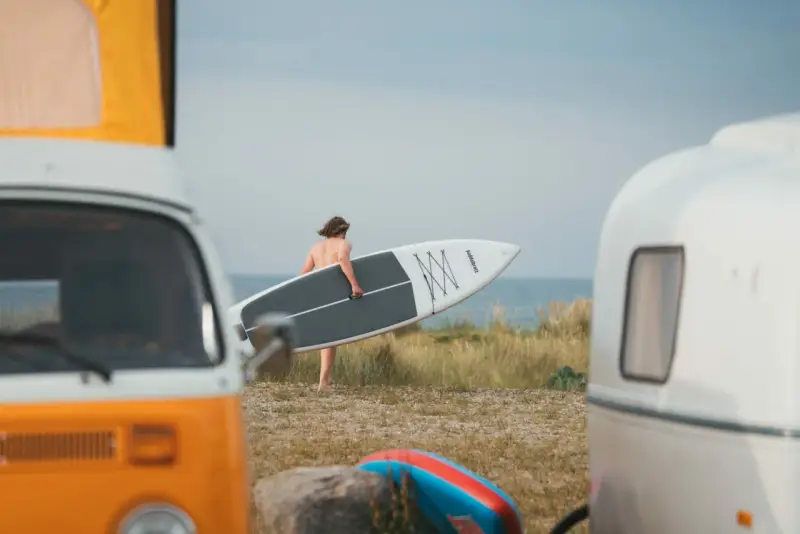 Mann mit SUP-Board am Strand von Fehmarn, bereit für die Wellen. Wohnmobile im Vordergrund.