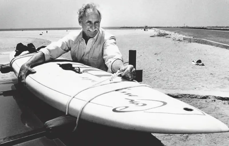 Jim Drake, ein Pionier des Surfens, lächelt stolz neben seinem Surfbrett am Strand.