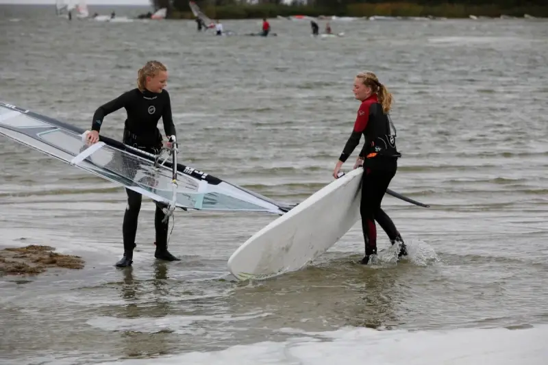 Zwei Mädchen bereiten sich auf das Windsurfen am IJsselmeer vor, tragen Neoprenanzüge und halten ihre Ausrüstung.